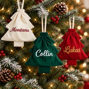 May include: Three tree-shaped fabric drawstring pouches hanging as ornaments. The cream pouch features the name Montana in red, the green pouch features the name Collin in white, and the red pouch features the name Lukas in gold script.