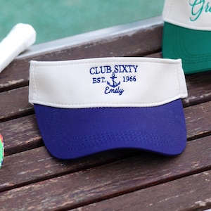 Può includere: Una visiera blu navy con un pannello frontale bianco caratterizzato da un ricamo blu scuro con la scritta CLUB SIXTY EST. 1966, un'icona a forma di ancora e il nome Emily sottostante. La visiera ha una tesa curva e una fascia bianca strutturata.