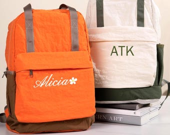Puede incluir: Dos mochilas con asas superiores y bolsillos delanteros con cremallera. La mochila izquierda es de color naranja brillante con detalles marrones y el nombre Alicia con un icono de flor. La derecha es blanca con detalles verde oscuro y las letras ATK.