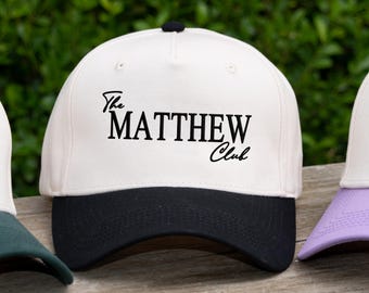 Puede incluir: Una colección de gorras de béisbol con coronas blancas y viseras de colores. Una gorra tiene una visera verde oscuro con el texto He's Golfing en verde oscuro. Otra gorra tiene una visera negra con el texto The MATTHEW Club en color negro.