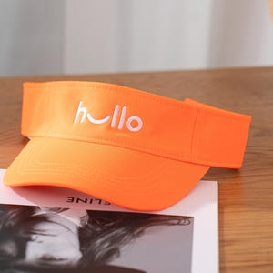 May include: Bright orange visor with the word "hello" embroidered in white. The visor is made of a sturdy fabric and has a curved brim. The visor is sitting on a magazine.