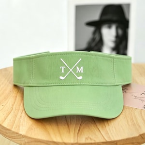 Pode incluir: Viseira verde claro com aba curva. A viseira apresenta as letras "T" e "M" com tacos de golfe cruzados em branco. A viseira está sobre uma superfície de madeira.
