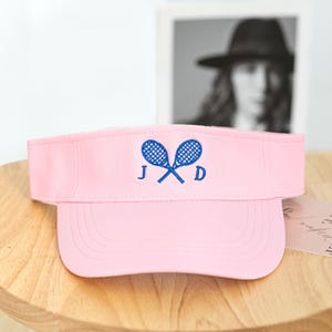 Può includere: Visiera da tennis rosa chiaro con visiera curva. La visiera presenta un design ricamato blu di racchette da tennis incrociate e le lettere "J" e "D". La visiera è esposta su una superficie di legno.