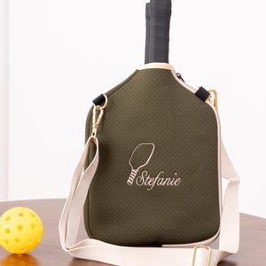 May include: Olive green pickleball paddle bag with a cream-colored strap and trim. The bag has a perforated texture and features an embroidered paddle design with the name "Stefanie". A yellow pickleball sits on a wooden surface.