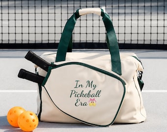 Puede incluir: Una bolsa de pickleball color crema con bordes y asas verde oscuro. El bolsillo frontal tiene el texto In My Pickleball Era con un pequeño lazo rosa y dos pelotas de pickleball amarillas. Dos palas negras están en el bolsillo lateral.