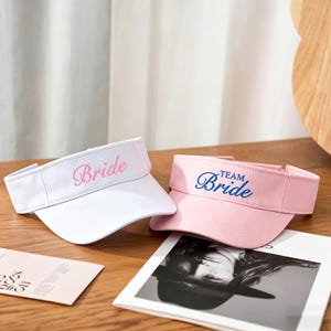 May include: Two visors, one white and one pink, are displayed on a wooden surface. The white visor has the word "Bride" in pink script, while the pink visor says "Team Bride" in blue script. The visors are likely for a bridal party.