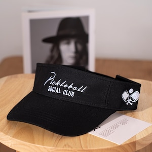 Könnte beinhalten: Schwarzes Visier mit dem weißen Schriftzug "Pickleball SOCIAL CLUB". Das Visier hat einen gebogenen Schirm und eine weiße Grafik von Pickleball-Schlägern und einem Ball. Das Visier liegt auf einer Holzoberfläche.
