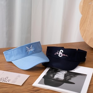 May include: Two visors, one light blue and one navy, are displayed on a wooden surface. The light blue visor has the name "Nicole" embroidered in white, with crossed baseball bats. The navy visor has the name "Jo 6son" embroidered in pink.