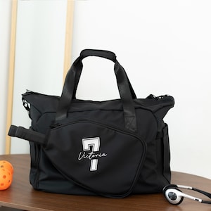 May include: Black duffel bag with a white graphic of a pickleball paddle and the name "Victoria". The bag has a top handle and shoulder straps. A pickleball paddle and headphones are next to the bag.
