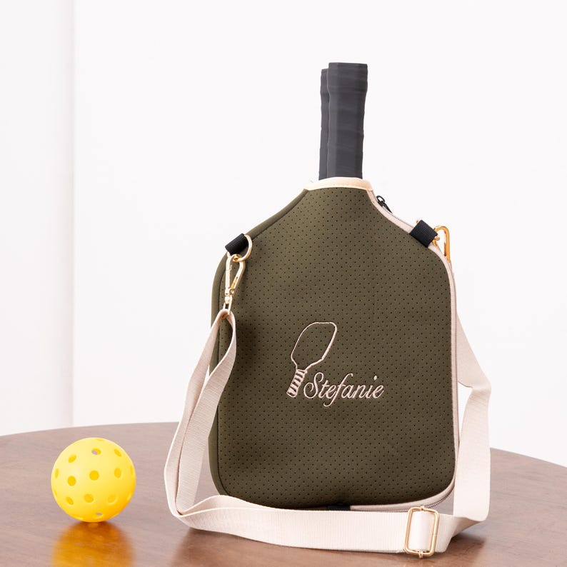 May include: Olive green pickleball paddle bag with a cream-colored strap and trim. The bag has a perforated texture and features an embroidered paddle design with the name "Stefanie". A yellow pickleball sits on a wooden surface.