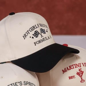 May include: A cream-coloured baseball cap with a black peak. The cap has embroidered text reading "HOT GIRLS WATCH FORMULA 1" and checkered flag graphics. Other caps with text are also visible.