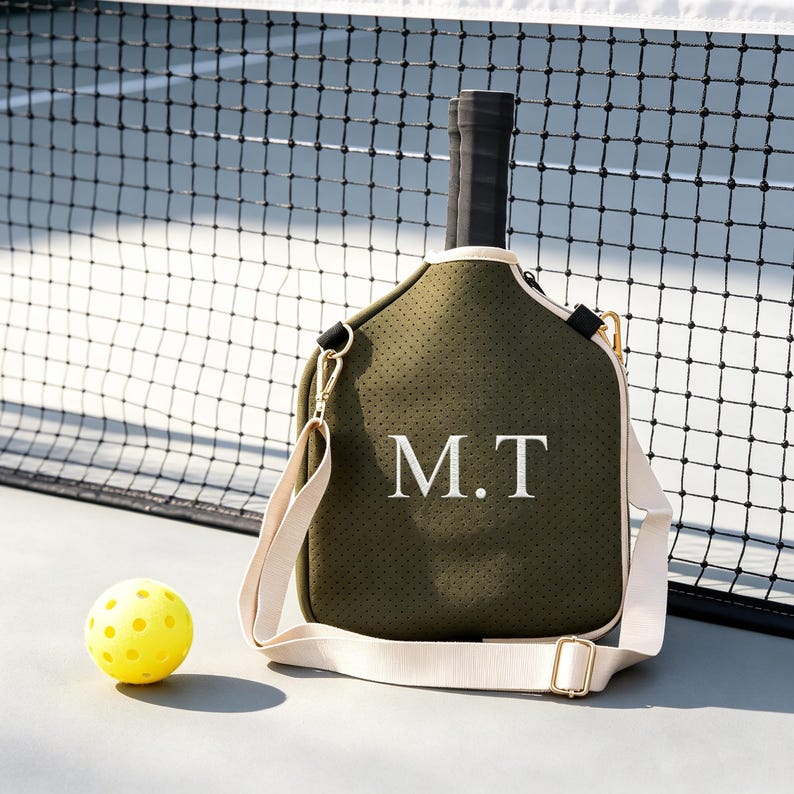 May include: Olive green pickleball paddle bag with white embroidered initials "M.T." Features a cream-colored shoulder strap and gold-colored hardware. A yellow pickleball sits on the court next to the bag. The background includes a black net.