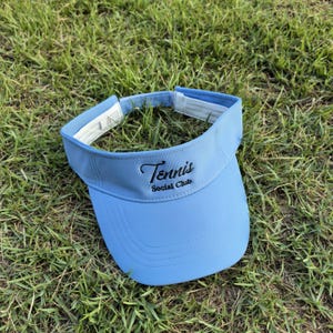 May include: A light blue visor featuring the embroidered black text Tennis Social Club on the front panel. The visor has a curved brim with stitched detailing and a white interior lining with an adjustable strap closure at the back.