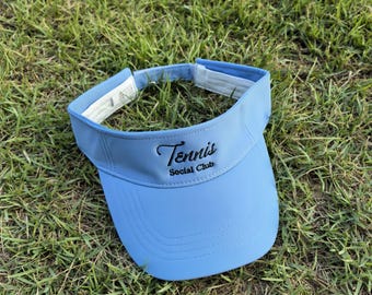May include: A light blue visor featuring the embroidered black text Tennis Social Club on the front panel. The visor has a curved brim with stitched detailing and a white interior lining with an adjustable strap closure at the back.