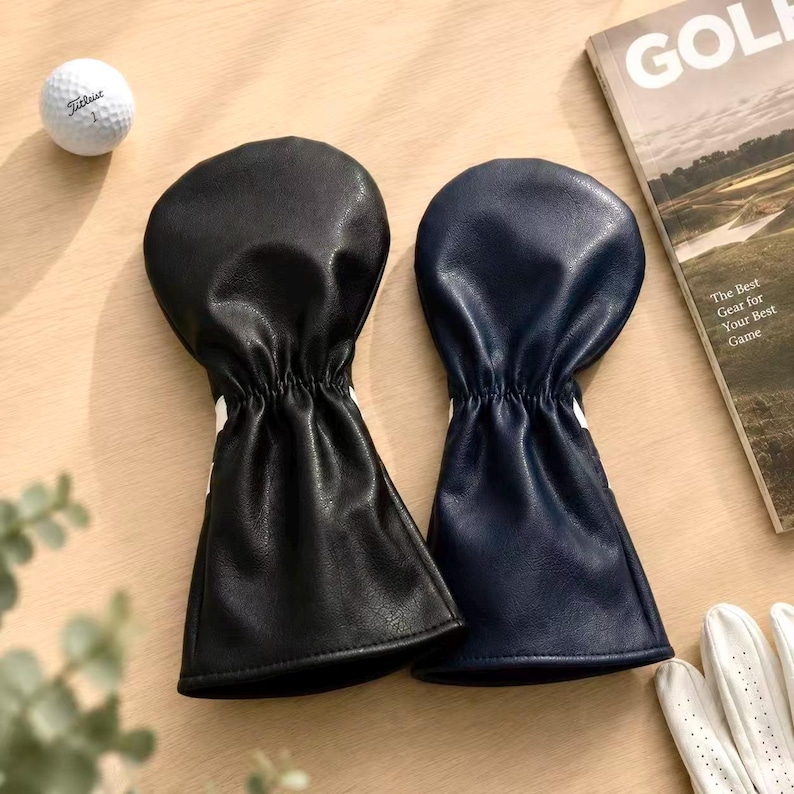 May include: Two golf club head covers, one black and one navy blue, with white accents. A golf ball and a magazine with the text "GOLF" are also visible. The magazine's cover reads "The Best Gear for Your Best Game."