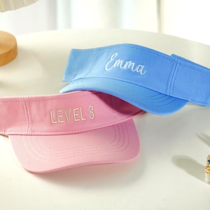 May include: Two visors, one light blue and one pink, are displayed. The blue visor has the name "Emma" embroidered in white script. The pink visor has "LEVEL 8" embroidered in white. Both visors are made of a soft fabric.