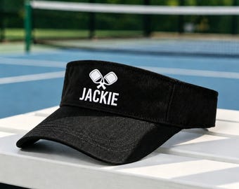May include: A black visor featuring an embroidered white graphic of two crossed pickleball paddles above the name JACKIE in white capital letters. The visor has a curved brim and a structured front panel with visible stitching details.
