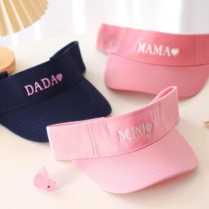 May include: Three visors in navy and pink. The navy visor has "DADA" embroidered in pink. The pink visors have "MAMA" and "MINI" embroidered in white. A cute accessory for sunny days.