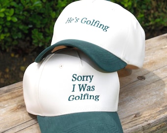 Puede incluir: Dos gorras de béisbol de color blanco roto con viseras de color verde oscuro. La gorra superior presenta el texto bordado "He's Golfing" en verde oscuro. La gorra inferior presenta el texto bordado "Sorry I Was Golfing" en verde oscuro.