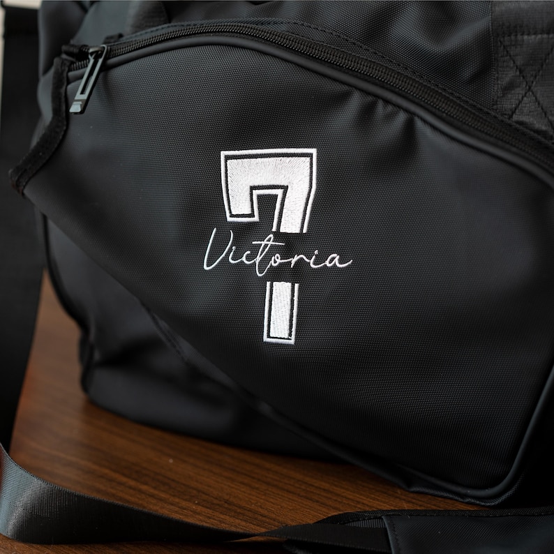 May include: Black duffel bag with a white embroidered design. The design includes the number 7, the name "Victoria", and a rectangular shape. The bag has a zipper closure and a black strap.