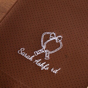 May include: Brown fabric with a dotted texture and a white embroidered design of two connected paddles with a bow. The text "Sarah Ashford rd" is embroidered below the paddles.