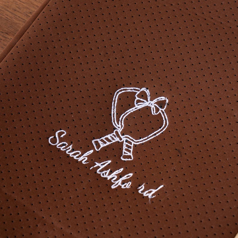 May include: Brown fabric with a dotted texture and a white embroidered design of two connected paddles with a bow. The text "Sarah Ashford rd" is embroidered below the paddles.