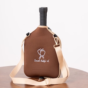 May include: Brown tennis racket cover with a beige shoulder strap and gold-colored hardware. The cover has a perforated texture and features an embroidered design with the text "Sarah Ashfords rd". The handle is black.
