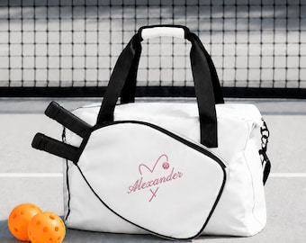 Puede incluir: Una bolsa de pickleball blanca con asas y bordes negros, que presenta un bolsillo frontal con forma de pala de pickleball. El bolsillo está bordado con el nombre Alexander en letra cursiva rosa, junto a un corazón rosa y una pequeña pelota rosa.