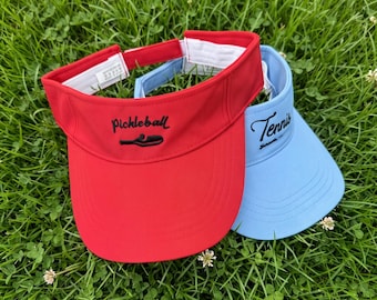 May include: A red visor with the word Pickleball and a paddle icon embroidered in black, and a light blue visor with the word Tennis embroidered in black. Both visors feature an adjustable white hook-and-loop strap closure at the back.
