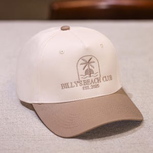 以下が含まれることがあります： 白のクラウンとベージュのつばが付いた野球帽。帽子には、ヤシの木と「BILLY'S BEACH CLUB EST. 2025」の文字が刺繍されたロゴが付いています。帽子には湾曲したつばと上部にボタンが付いています。