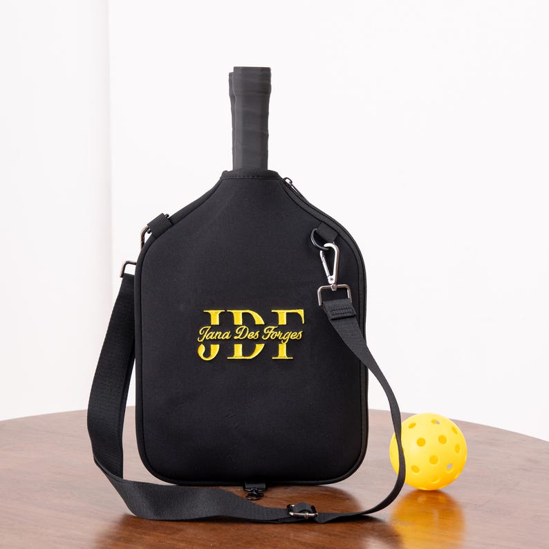 May include: Black pickleball paddle cover with a shoulder strap and a yellow pickleball. The cover has the text "Jana Des Forges" and "JDF" embroidered in gold. The paddle handle is visible.