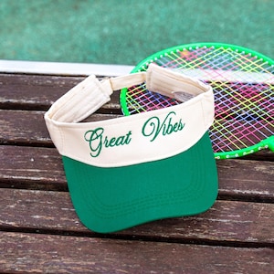 以下が含まれることがあります： 前面に緑色の筆記体で「Great Vibes」と刺繍された、クリーム色の本体と緑色のつばを持つサンバイザー。サンバイザーには背面に調節可能なストラップが付いており、カラフルなガットが張られた緑色のテニスラケットの前に置かれています。