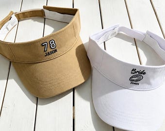 May include: A tan visor with the embroidered text 78 JASON in black and white, and a white visor with the embroidered text Bride Team and a small circular graphic in black. Both visors feature an adjustable strap with a white interior lining.