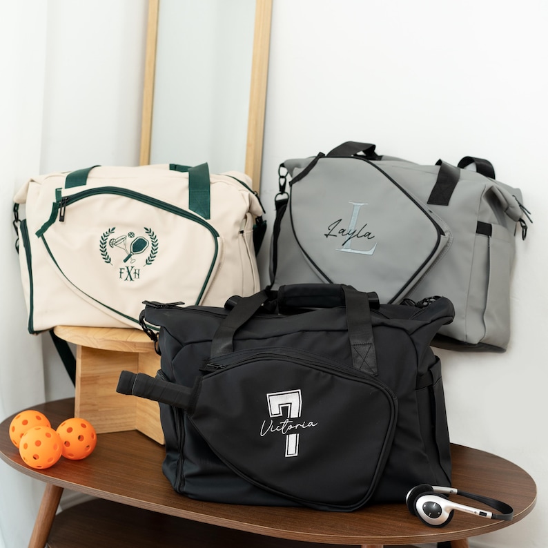May include: Three personalized pickleball bags in cream, gray, and black. The cream bag has green accents and a pickleball emblem. The gray bag features a name in script. The black bag has a pickleball paddle design with a name. Orange pickleballs and headphones are also visible.