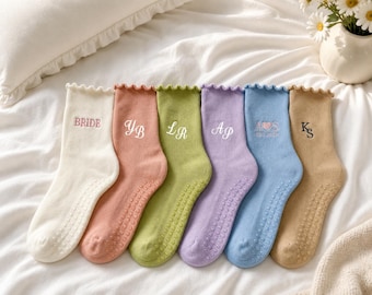 May include: Six pairs of ankle socks with ruffled cuffs and non-slip grips on the soles. The socks are cream, peach, olive green, lavender, light blue, and tan. Each pair features embroidered text or initials: BRIDE, YB, LR, AP, A heart S, and KS.