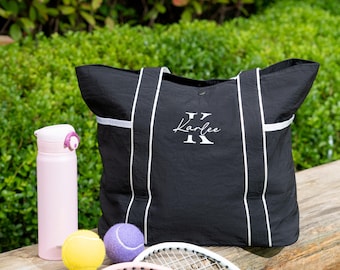 Puede incluir: Un bolso tote negro con ribetes blancos y el nombre Karlee bordado en blanco sobre una letra K grande, una botella de agua rosa claro, una pelota de tenis amarilla, una pelota de tenis morada y dos raquetas de tenis con marcos blancos y detalles rosas.