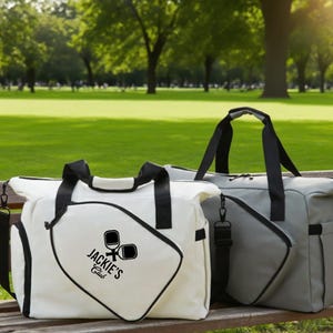 May include: Two duffel bags, one white and one gray, with black handles and straps, sit on a wooden bench. The white bag features a logo that reads "Jackie's Club" with pickleball paddles. The bags are in a park setting.