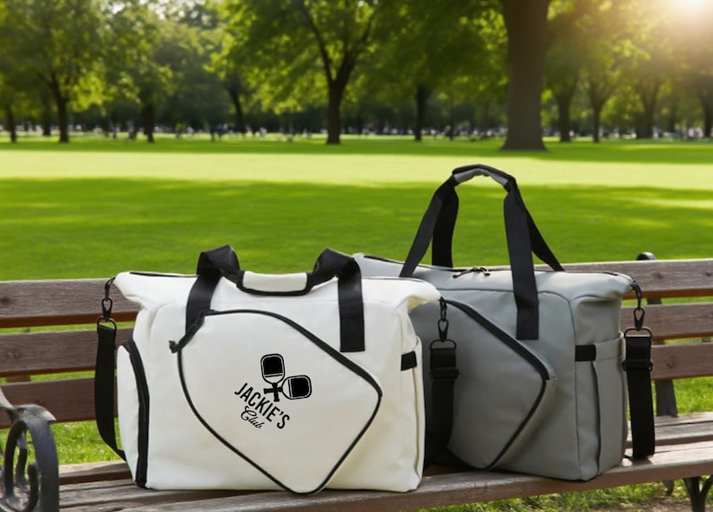 May include: Two duffel bags, one white and one gray, with black handles and straps, sit on a wooden bench. The white bag features a logo that reads "Jackie's Club" with pickleball paddles. The bags are in a park setting.