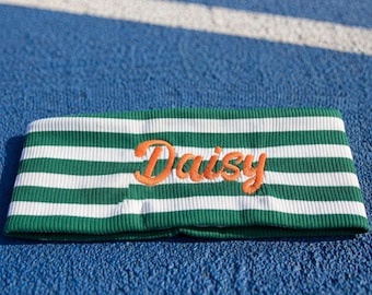 Puede incluir: Una cinta para el pelo acanalada con rayas horizontales verdes y blancas. La palabra Daisy está bordada en letra cursiva naranja en el centro de la tela. El accesorio tiene una apariencia texturizada y elástica con un patrón de rayas uniforme.