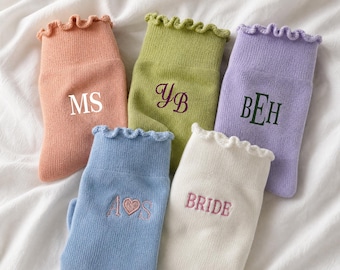 May include: Five pairs of ruffled-top socks in peach, lime green, lavender, light blue, and cream. The peach sock features white text MS, the lime green has dark purple text YB, the lavender has dark green text BEH, the blue has pink text A heart S, and the cream has pink text BRIDE.