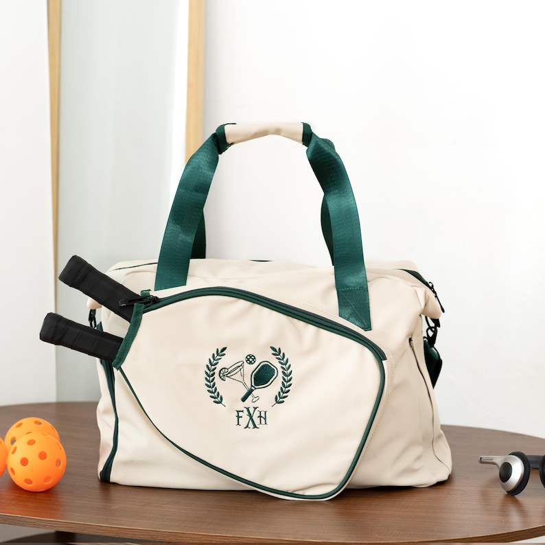 May include: Cream-colored pickleball bag with green trim and handles. The bag features a green embroidered design of a pickleball paddle, ball, and laurel wreath. Two black pickleball paddles are visible, along with orange pickleballs.