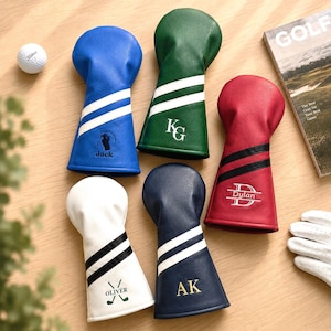 May include: A collection of personalized golf club head covers in various colors, including blue, green, red, white, and navy. Each cover features a unique design with initials or names, along with white stripes. A golf ball, a golf glove, and a golf magazine are also visible.