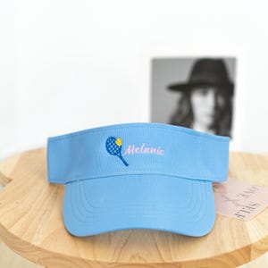 May include: Light blue visor with a tennis racket and ball embroidered on the front. The name "Melanie" is embroidered in pink script. The visor is made of a soft material and has a curved brim.