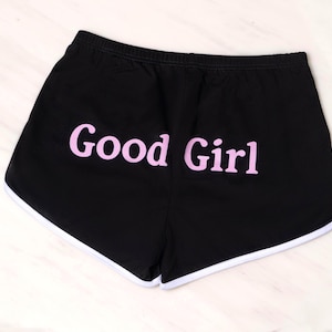 Peut inclure: Short de sport noir avec un liseré blanc le long de l'ourlet incurvé et le texte Good Girl imprimé en lettres rose clair au dos. Le short est doté d'une taille élastique et présente une conception simple et fonctionnelle.