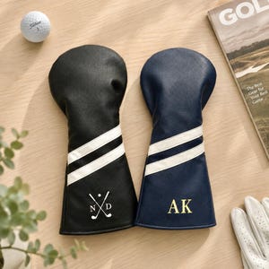 May include: Two leather golf club head covers, one black and one navy blue, each with white stripes and personalized initials. A golf ball and a golf magazine are also visible.