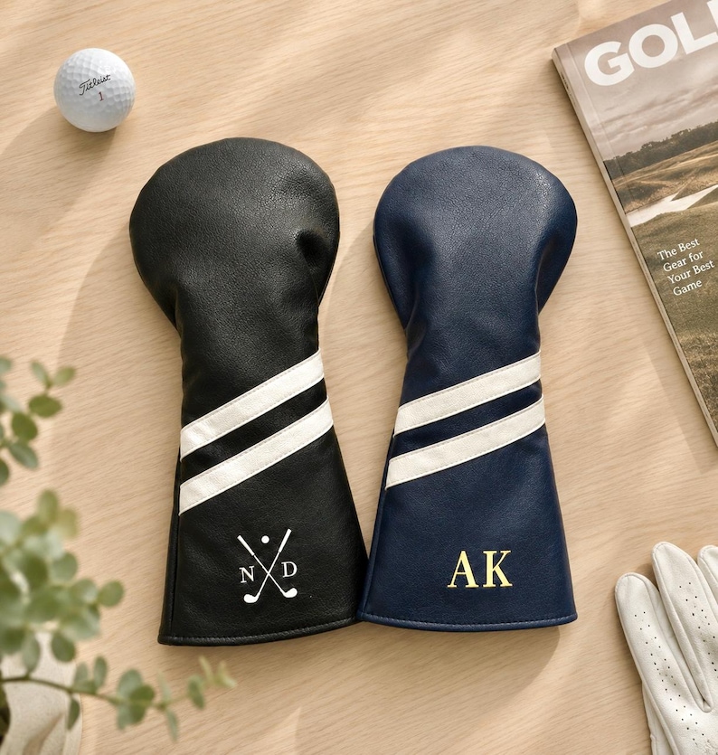 May include: Two leather golf club head covers, one black and one navy blue, each with white stripes and personalized initials. A golf ball and a golf magazine are also visible.