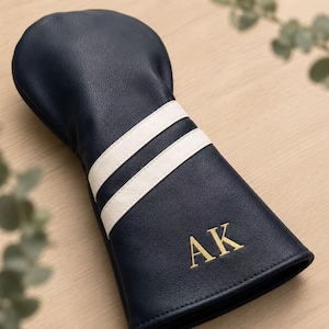 May include: Navy blue golf club head cover with two white stripes and gold embroidered initials "AK". The cover is made of a leather-like material and is designed to protect golf clubs.