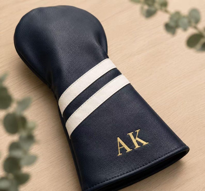 May include: Navy blue golf club head cover with two white stripes and gold embroidered initials "AK". The cover is made of a leather-like material and is designed to protect golf clubs.