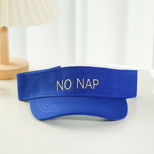Pode incluir: Uma viseira azul com o texto "NO NAP" bordado em dourado. A viseira tem uma aba curva e uma faixa de tecido. A viseira está sobre uma superfície branca.