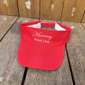 Op de afbeelding: Een felrode zonneklep met de witte geborduurde tekst Morning Ritual Club op het voorpaneel. De klep heeft een gebogen rand met zichtbare stiksels en een verstelbare band aan de achterkant met een witte binnenvoering.